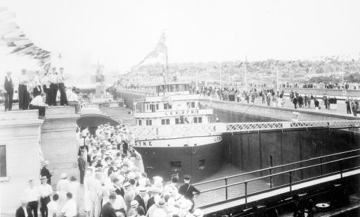 Photo en noir et blanc d’une foule rassemblée autour d’une écluse à l’intérieur de laquelle se trouve un bateau pavoisé.