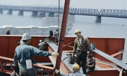 Men in hardhats work on an orange metal structure above the St. Lawrence.