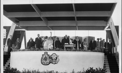 Des invités d’honneur et des officiers de la marine sont sur une estrade arborant les armoiries du Canada et des États-Unis.