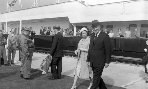 En avant-plan, on voit la reine et le président qui marchent et, à l’arrière-plan, un navire avec des passagers à bord.