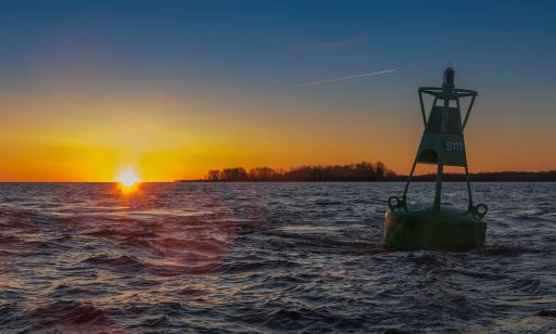 Close-up of a large green buoy bearing the inscription S111. On the horizon, an island is silhouetted against the sunrise.