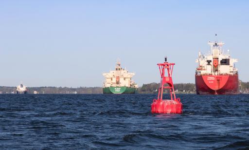 Photo taken from a boat on the river showing a close-up of a red buoy and a rear view of cargo ships in the background.