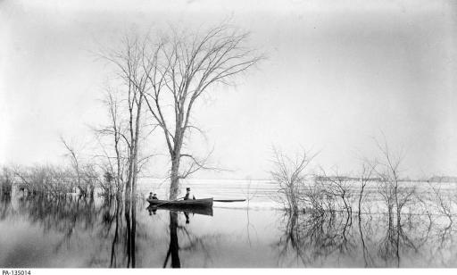 Black and white photo of a man rowing two children in a boat past a row of trees in the water.