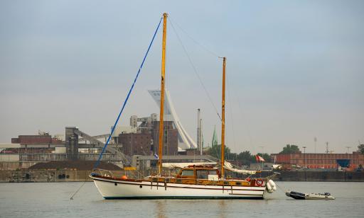 A two-masted blue and white wooden sailboat anchored on the river, with an inflatable boat tied behind it.