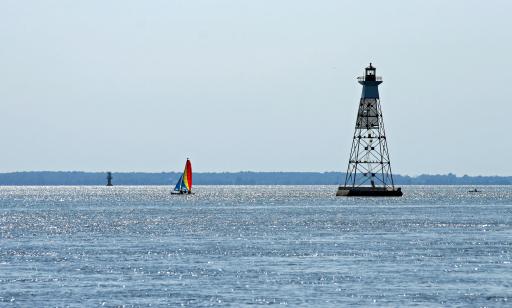 Un petit voilier à un mât arborant une voile rouge, jaune et bleu et un foc bleu navigue sur le fleuve entre deux phares.