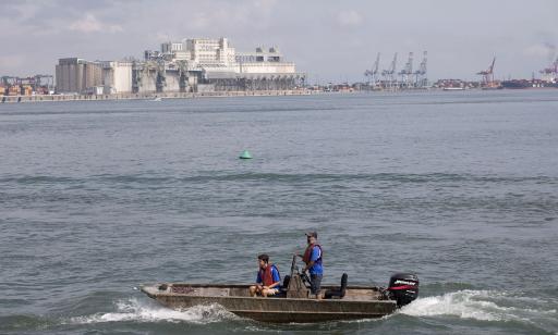 Two men in a small motorboat, with major port facilities in the distance.