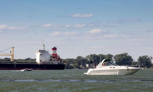 On voit des habitations sur une rive boisée devant laquelle se déplacent deux bateaux à moteur près d’un immense cargo.