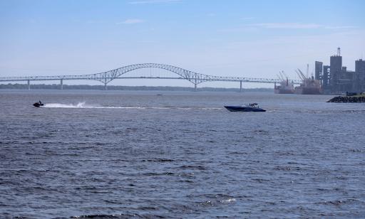 A personal watercraft passes near a motorboat, throwing up a large wake. Two large cargo ships are moored in the port.