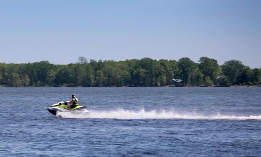 A man in a fast-moving personal watercraft leaves a trail of foam on the river, in front of a wooded shoreline.