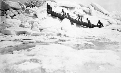 Black and white photo of six men hoisting a canoe onto a large mound of ice next to the frozen water.