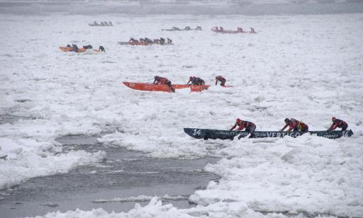 Around 30 men push seven canoes of various colours over a wide field of ice floating on the river.