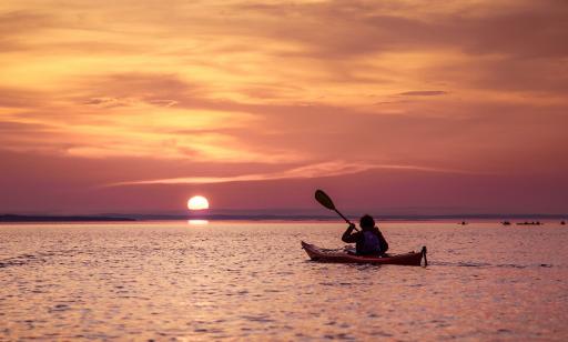 Un homme se déplace en direction du soleil couchant sous un ciel orangé en pagayant dans un kayak sur une eau assez calme.