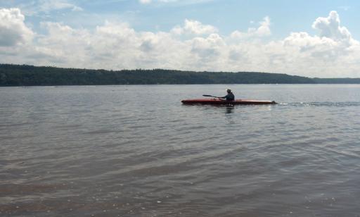 A man paddles a kayak on calm water during the summer. A wooded shoreline stands out against the horizon.