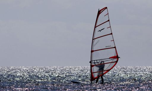 Un homme navigue à bord d’une planche à voile sur l’eau miroitante du fleuve durant une journée ensoleillée d’été.