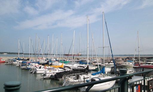 Numerous sailboats and motorboats moored at a dock. Two cargo ships and a small sailboat can be seen in the distance.