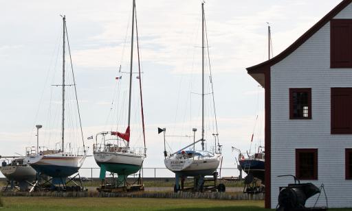 Cinq voiliers reposent sur leurs remorques à gauche d’un bâtiment ancien en bois blanc aux fenêtres d’un rouge foncé.