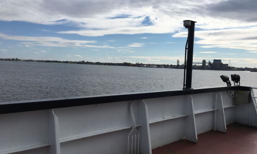 From behind the guardrail of a ferry, a boat on the river can be seen in the distance, with port facilities behind it.