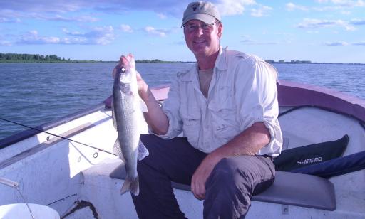 On a sunny day, a man seated in a boat on the river holds up a large fish.