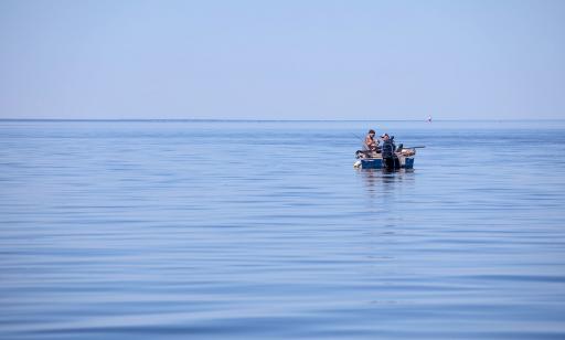 Two men fish from a boat in the middle of a wide stretch of water. A lighthouse can be seen in the distance.