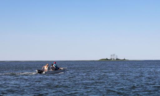 Three men drive a motorboat toward a tiny island with a few trees and shrubs on it.