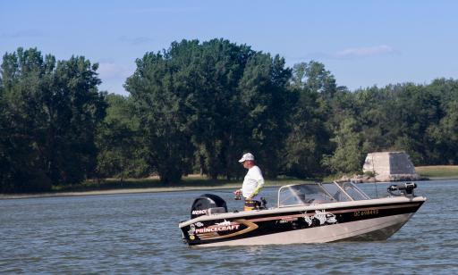 A man holding a fishing rod stands in a motorboat in front of a tree-covered island.