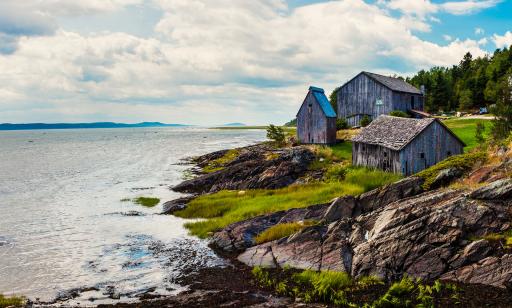 Three unpainted grey wooden buildings on a rocky shore. The smallest one is narrow and has a steeply pointed roof.