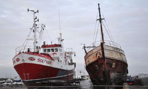 Two boats out of water. One has a sound metal hull, while the other has a severely damaged wooden haul.