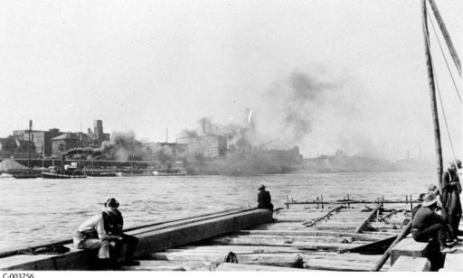 Men seated on a timber raft. In the distance, the city can be seen through clouds of smoke emitted by boats.