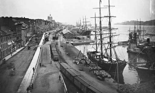 Des barils et du bois sont empilés sur le quai près des grands voiliers. Un train et des calèches se déplacent non loin.