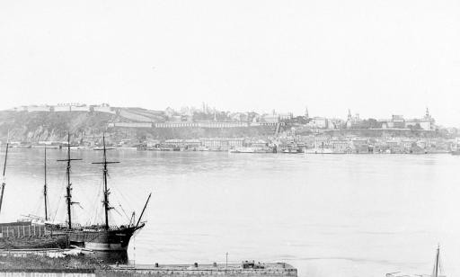 A docked sailing ship. Buildings, churches and ramparts can be seen on a headland on the far shore.