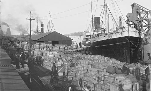 Men on a wharf, where many wooden boxes and barrels are piled up next to the steamships.