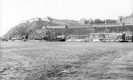 Distant view of steamships at the wharf, ramparts, a rocky headland and city buildings.