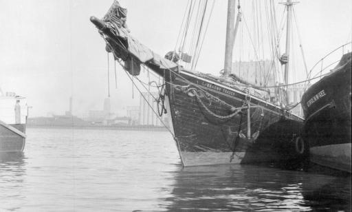 Un voilier en bois aux voiles ferlées côtoie de près un autre bateau au quai. Au loin, on voit des installations portuaires.