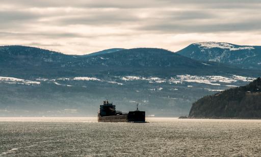 A ship with a black hull sails along a river past majestic snow-covered mountains.