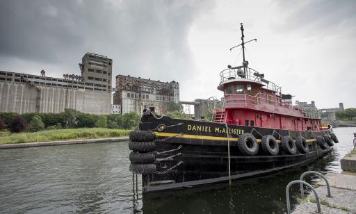 A boat with old tires on its sides as fenders moored in a basin. Derelict industrial buildings can be seen in the background.