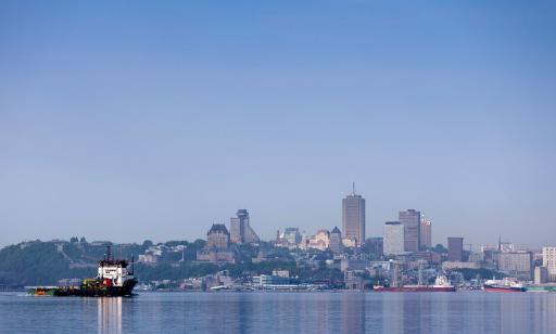 A black and white boat on the river. In the distance, the city and cargo ships by the port can be seen.