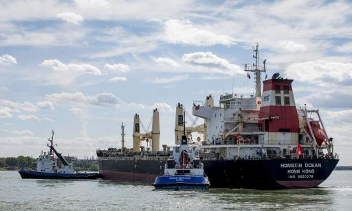 Two sturdy blue and white boats tow a large ship along the river. Lifting gear can be seen on the deck.