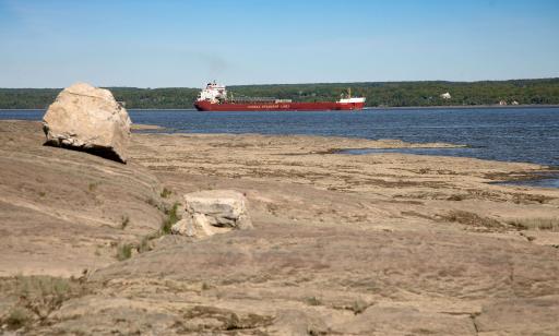 View from the rocky shore of a cargo ship leaving a wake behind it. The other shore is forested.