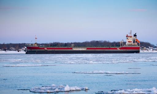 Un navire gris, noir et rouge se déplace sur l’eau où flottent des glaces non loin de la rive enneigée où il y a des maisons.