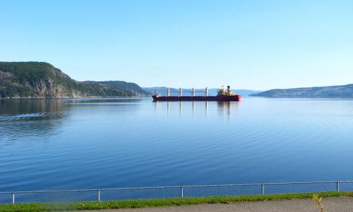 A red and white cargo ship with four lifting devices on its deck sits on the calm water of a bay surrounded by mountains.