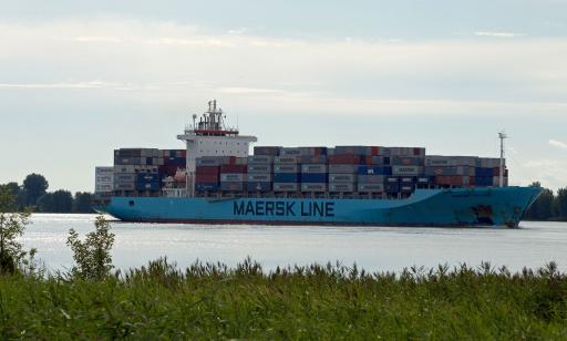 View from the shore of an enormous ship, heavily loaded with containers. Its pale blue hull is rusted at the bow.