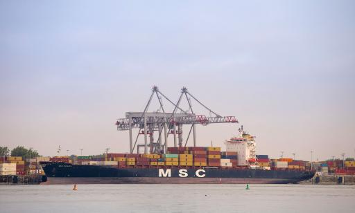 On voit les grandes flèches de deux grues-portiques au-dessus d’un navire imposant qui est lourdement chargé de conteneurs.