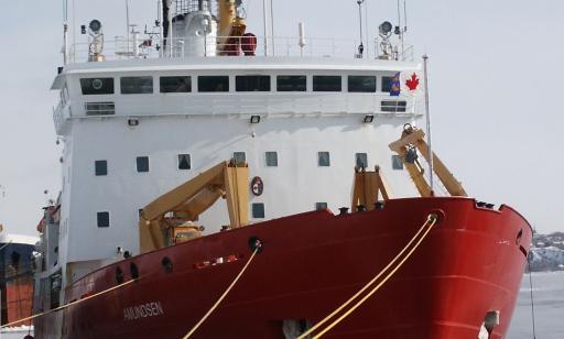 A red and white ship tied up at the wharf with several cables, surrounded by floating ice.