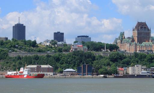 A red and white ship on the water. Buildings, trees, an escarpment and ramparts can be seen on the headland behind it.