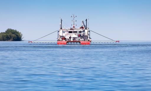 Des sondes accrochées à deux longs tuyaux de chaque côté d’un navire rouge et blanc sont plongées sous la surface de l’eau.