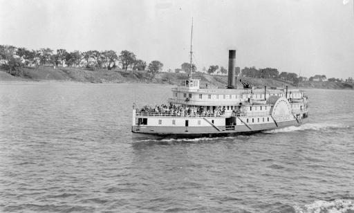 Une photo en noir et blanc montre un bateau, doté d’une cheminée et de deux roues à aubes, et au loin la rive d’une île.