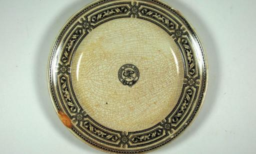 White plate with a crackled yellow glaze, bearing a company logo in the centre and blue decorative motifs around the edge.