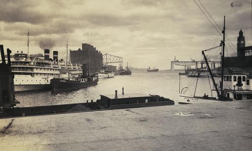 View from a wharf of steamboats and, in the distance, cranes mounted on a bridge with a missing span.