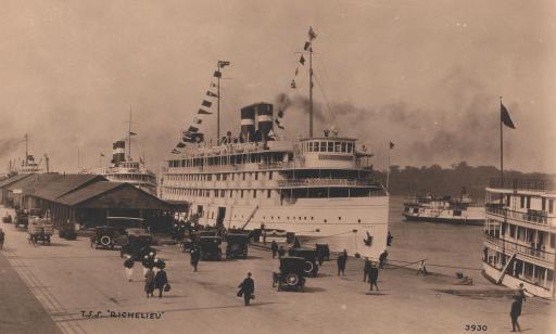 A white boat decked in flags with smoke rising from its stack. Strollers, cars and buildings can be seen on the wharf.