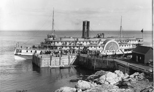 On a wharf, horse-drawn carriages and a crowd of people gather near a boat with many passengers on board.